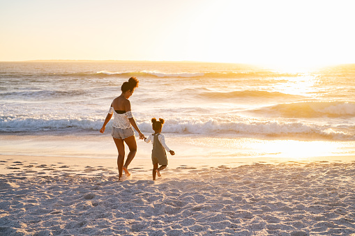 Happy african american mother relaxing with cute little girl at beach while walking towards the sea. Black family walking towards ocean on beach during sunset with copy space. Mother and child running barefoot on seaside while holding hands.