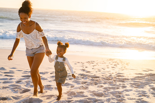 Young beautiful woman with her adorable little cute girl running on tropical beach during sunset. Cheerful and cute african american daughter walking and playing with mother on beach together. Lovely bigger sister and female child enjoying beach with copy space.