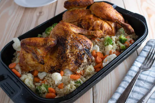 Delicious and healthy homemade family meal with roasted whole chicken, brown basmati rice and imperial vegetables. Served in a roasting pan on wooden table. Ready to eat