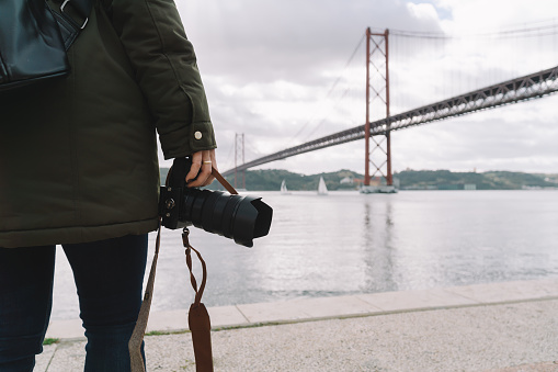 Unrecognizable young woman holding camera in close-up with famous Lisbon bridge in background