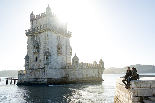 Couple in love sitting on a wall while talking to each other with the belem tower on the side, on a sunny morning.