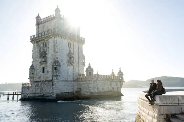 Photo of Young woman and man sitting next to the Belem Tower in Lisbon.