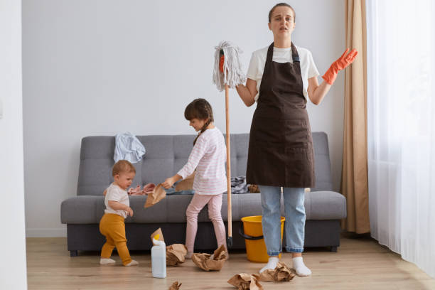 foto interior de una mujer joven vestida con camiseta blanca, delantal marrón y jeans, limpiando la casa, posando con sus hijos, sosteniendo la fregona en las manos, teniendo una expresión facial impotente. - 6 11 meses fotografías e imágenes de stock