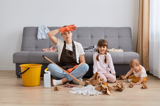 Exhausted Caucasian woman wearing white t shirt, brown apron and jeans, cleaning house, posing with her children, keeping hand on her forehead, being tired long hours doing house hold chores.