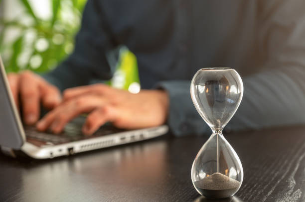 Hourglass with time running out in an office as a symbol of time pressure at work in the background a man in a shirt at his laptop, in the foreground running hourglass hourglass stock pictures, royalty-free photos & images