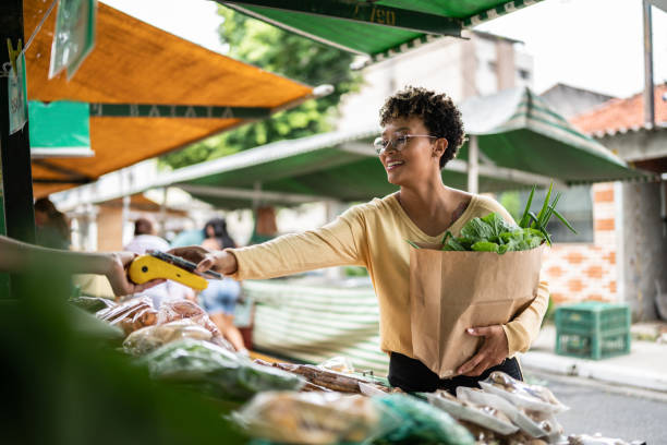 jovem pagando com celular em um mercado de rua - groceries women bag customer - fotografias e filmes do acervo