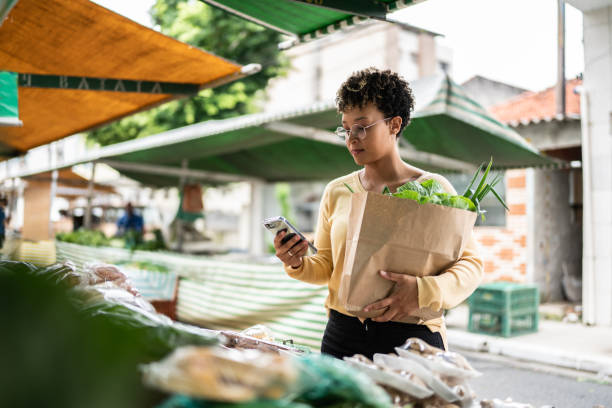 une jeune femme utilisant le téléphone portable dans un marché de rue - organic farmers market market vegetable photos et images de collection