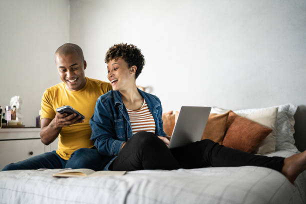 el esposo mostrando algo en el teléfono móvil a su esposa en casa - animando hombre con ordenador portátil en la cama fotografías e imágenes de stock