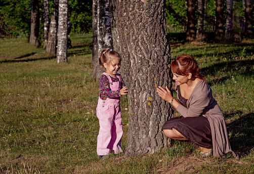 Middle aged woman playing with little girl hiding behind tree in summertime