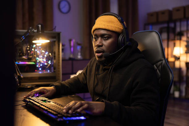 Shocked male professional computer gamer opens mouth looking at computer screen, talks to team through headset, presses buttons of backlit keyboard. Dark room lit by purple led light