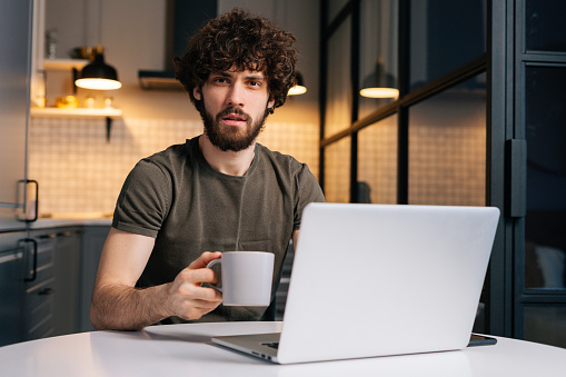 Portrait of confused curly young freelancer male holding in hand cup with morning coffee sitting at table with laptop computer, looking at camera, in kitchen with modern interior, remote home office.
