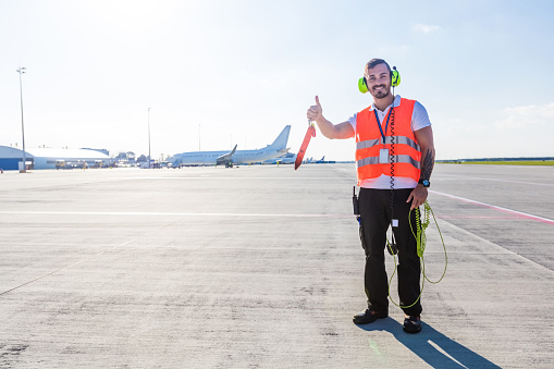 Outdoor shot of young man standing outside in front of airplane and raising hand with thumb up. Airport ground crew at work.