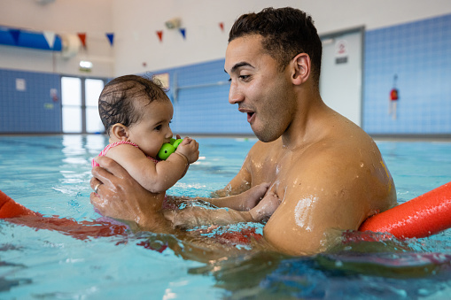 A man holding and swimming with his baby using a woggle during a swimming class in Boldon, North East England. The man is looking excitedly at his baby while she sucks on a toy.