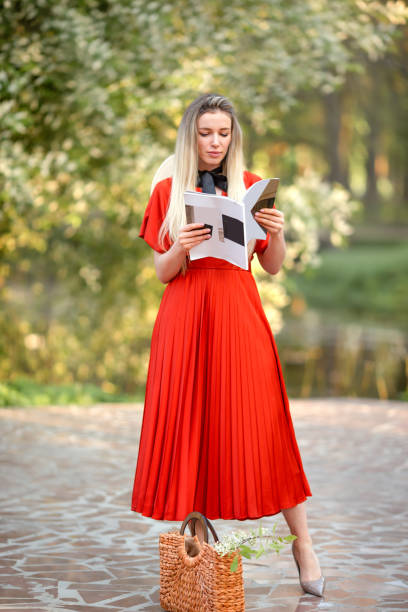 Beautiful girl in a red dress with a straw hat in a blooming park reads a magazine