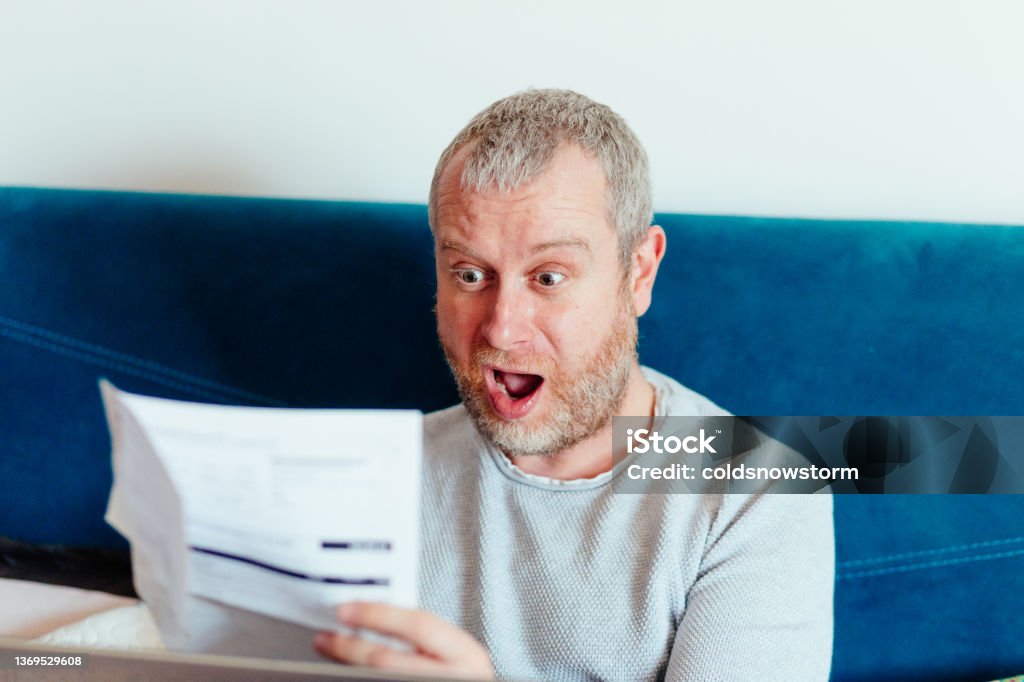Worried man checking bills at home Portrait of a mid adult man checking his energy bills at home. He has a worried expression and touches his face with his hand while looking at the bills. Focus on the man while the interior architecture of the house is defocused. Mortgage Loan Stock Photo