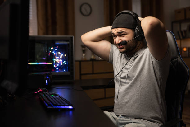 A man playing professionally on a computer with a headset puts his hands behind his head in anger, disappointment, lost a round in a video game, a middle-aged guy in a dark room
