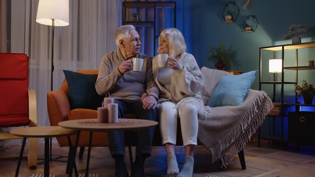 Carefree mature family couple relaxing on sofa at home, enjoying conversation with cups of hot tea