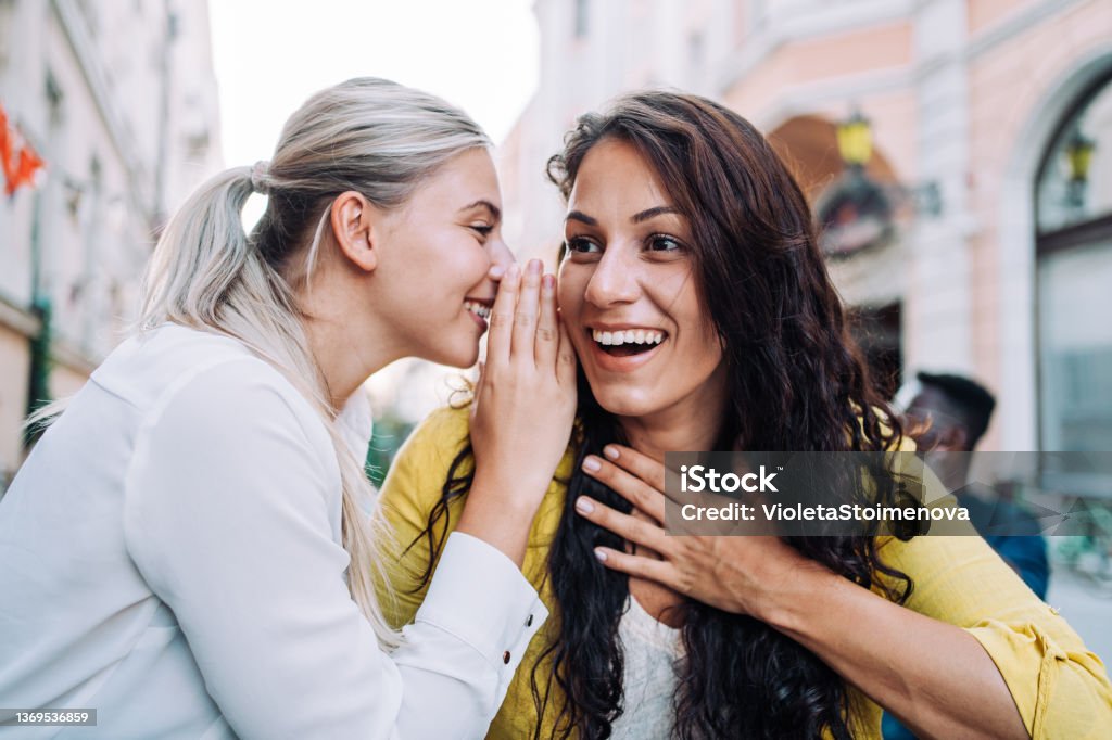Two female young friends sitting in city cafe and gossiping. Shot of young happy women talking and laughing while drinking coffee together in a sidewalk cafe. Whispering Stock Photo