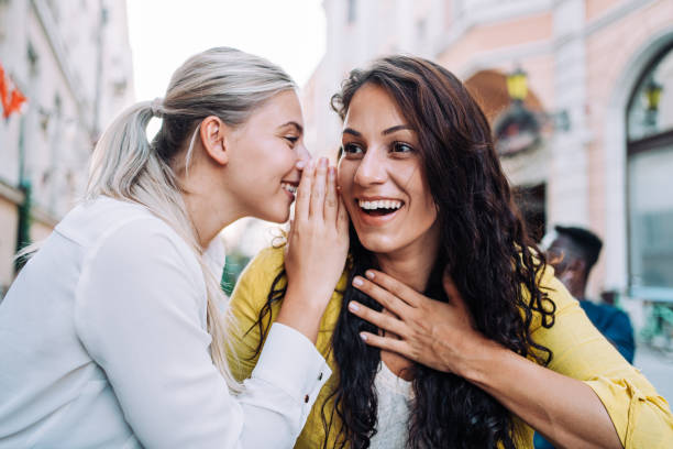 Two female young friends sitting in city cafe and gossiping. Shot of young happy women talking and laughing while drinking coffee together in a sidewalk cafe. whisperer pictures stock pictures, royalty-free photos & images
