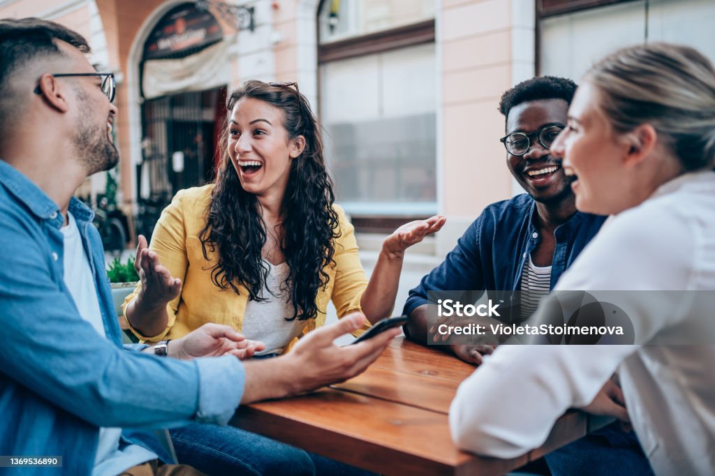 Young friends enjoying a coffee together. Shot of multiracial group of young people sitting in a sidewalk cafe talking, drinking coffee, smiling and having fun. Friendship Stock Photo