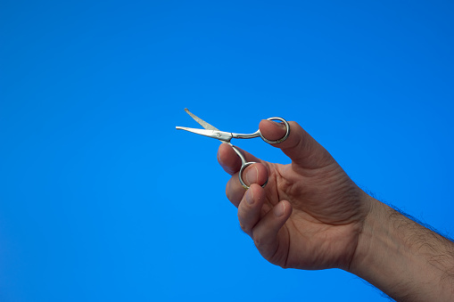 Small all metal scissor held in use by Caucasian male hand. Close up studio shot, isolated on blue background.