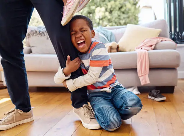 Photo of Shot of a little boy throwing a tantrum while holding his parent's leg at home