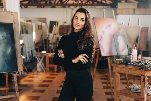 Confident painter standing in her workshop Confident female painter standing in her workshop with her artwork behind her. Young professional artist looking at the camera while standing with her arms crossed. artists stock pictures, royalty-free photos & images
