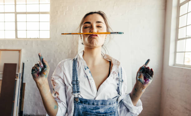 Playful female painter making a funny face with a paintbrush. Young female artist standing in her art studio with her blank canvas in the background. Creative young woman having fun in her atelier.