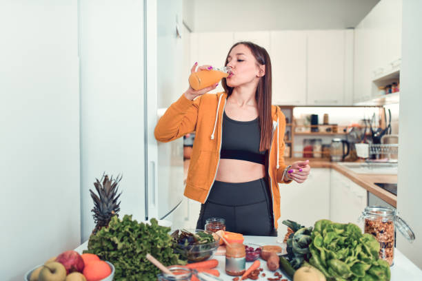 weiblicher trink-smoothie, um eine gesunde ernährung zu beginnen, während sie den hauptgang zu hause zubereitet - juicer food and drink healthy eating sport stock-fotos und bilder