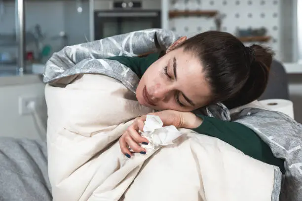 Young ill woman wrapped in blanker blowing her nose in to the toilet paper. Sick female sitting at home on sofa try to warm up suffering from common cold or flu, feeling exhausted and tired.