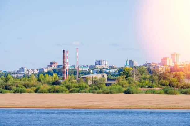 View of the urban industrial area across the river. Summer green country landscape with factory pipes.