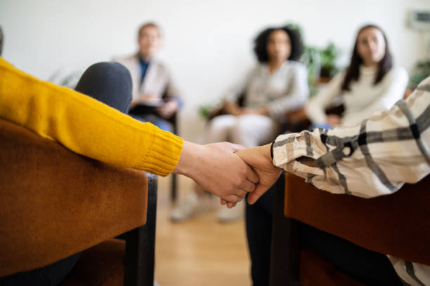 Holding hands for comfort during a support meeting Two people supporting each other by holding hands during a therapy meeting, the rest of the group are slightly out of focus nervous holding hands stock pictures, royalty-free photos & images