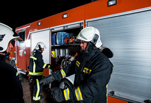 Two firefighters at the fire station, one firefighter using holding a fire hose while his colleague is storing a chainsaw inside of a fire truck equipment compartment