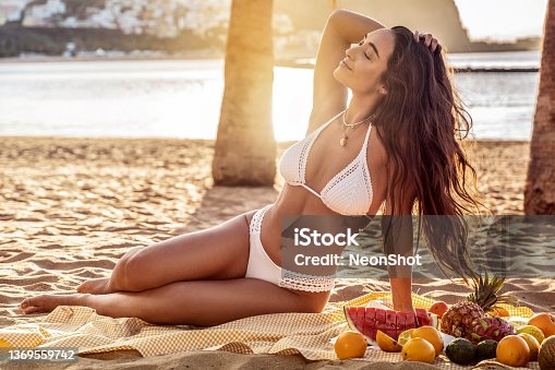 istock Young woman with beautiful fit tan body having picnic with fresh fruits at the empty tropical beach. Girl with long dark hair and natural look wearing white fashionable bikini. 1369559742