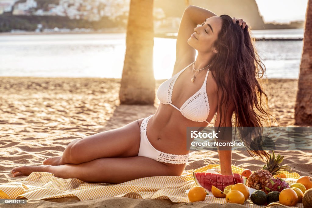 Young woman with beautiful fit tan body having picnic with fresh fruits at the empty tropical beach. Girl with long dark hair and natural look wearing white fashionable bikini. Young woman with beautiful fit tan body having picnic with fresh fruits at the empty tropical beach. Girl with long dark hair and natural look wearing white fashionable bikini. Vacation, day off or weekend activities concept. Real people lifestyle and emotions. Women Stock Photo