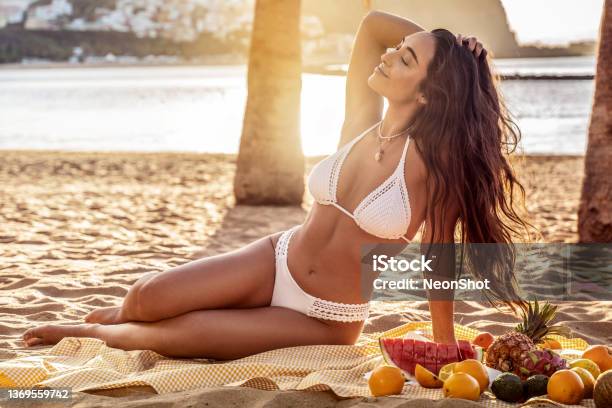 Young Woman With Beautiful Fit Tan Body Having Picnic With Fresh Fruits At The Empty Tropical Beach Girl With Long Dark Hair And Natural Look Wearing White Fashionable Bikini Stock Photo - Download Image Now