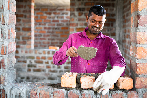 Happy smiling construction worker constructing or building wall by placing bricks and cement - concept of hard working, manual labor and daily wagers lifestyle.