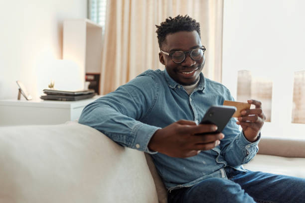 Making some quick payments online. Shot of a young man holding his credit card while using a smartphone at home. Cheerful young man doing online shopping while using his cellphone and credit card inside of an apartment during the day. person paying stock pictures, royalty-free photos & images
