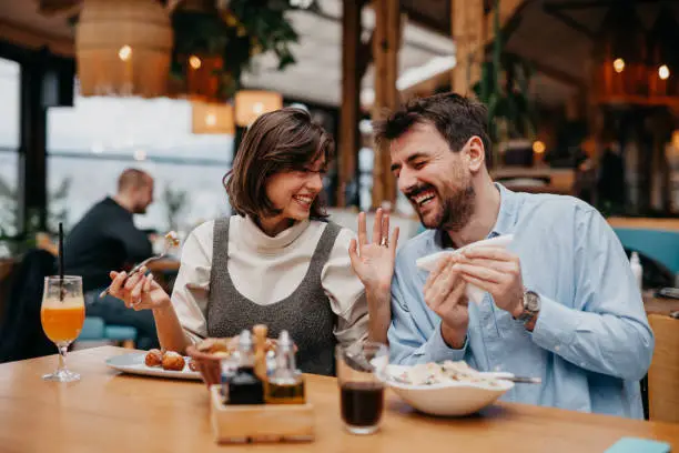 Two Young Friends Meeting For Drinks And Food In Restaurant, laughing, and eating.