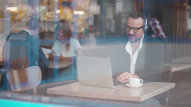 Ver a través de la ventana de un hombre de negocios con auriculares que tiene una videollamada en la computadora portátil en la cafetería. En tiempo real. photo