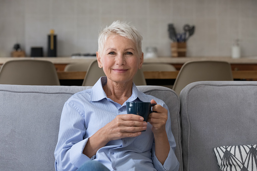 Happy calm carefree middle aged lady enjoying good morning, drinking hot fresh tasty coffee at home, relaxing on comfortable couch, holding ceramic mug, cup of tea, smiling. Head shot portrait