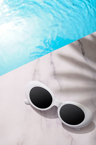 Pair of retro vintage sunglasses sitting poolside with the sun reflecting in the water and the shadow of a palm tree with copy space and room for text