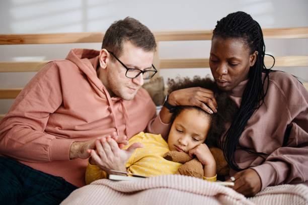 menina doente abraça com a mãe e o pai na cama enquanto eles lêem uma história para ela - cold and flu flu virus bed couple - fotografias e filmes do acervo