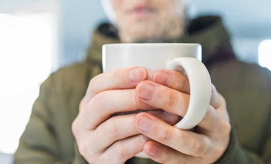 Closeup hands holding a white mug of hot tea with a blurry young man in a green winter topcoat a bright and soft background, selective focus.