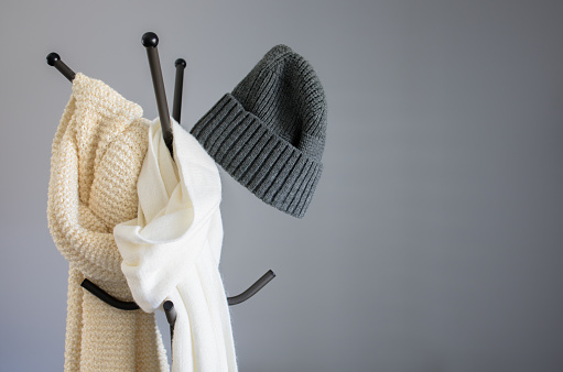 Winter garment accessories, minimal style of white cotton scarf, soft knitted scarf, and casual knitted hat hanging on a coat hook stand against the wall backgrounds.