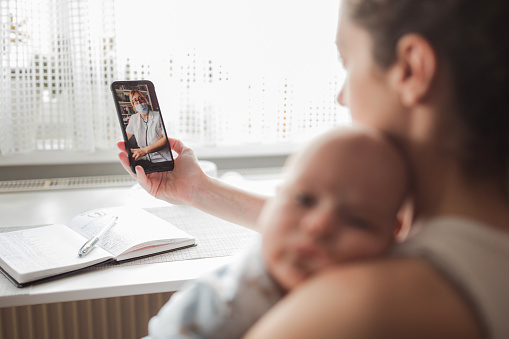 Young woman with a newborn baby on her shoulder holding a smart phone and calling a doctor via video call