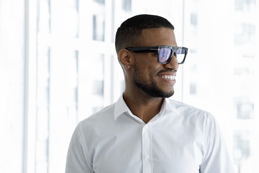 Happy young millennial Black businessman in formal white shirt and glasses thinking of future success, startup growth, standing at office window, looking away, smiling. Head shot portrait