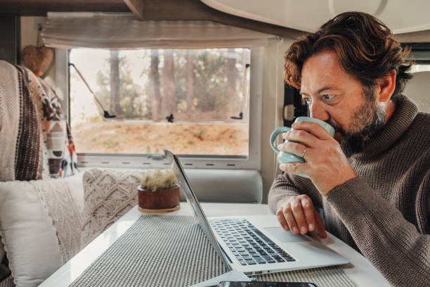 Mature man working on laptop computer inside a camper van with nature outdoors view outside the window. Concept of freedom and vanlife lifestyle. Smart remote working  online Mature man working on laptop computer inside a camper van with nature outdoors view outside the window. Concept of freedom and vanlife lifestyle. Smart remote working  online digital nomad stock pictures, royalty-free photos & images