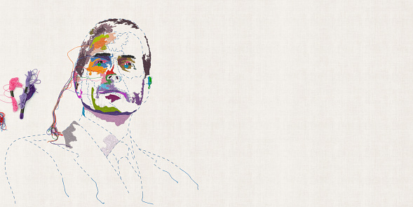 An overhead view of a embroidered and sewn portrait of a male head looking towards the camera, on plain off-white flat fabric. Multicoloured patches have been embroidered and loose threads are left hanging and intertwined to the side. a few skeins of thread are visible at the wide.