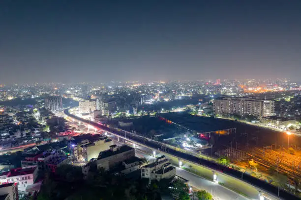 aerial drone shot showing elevated metro train station and tracks over busy street with light trails from traffic and cityscape in gurgaon, jaipur India..
